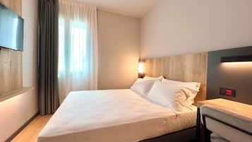 A hotel room with a bed, a TV mounted on the wall, a bedside table, and a window with curtains.
