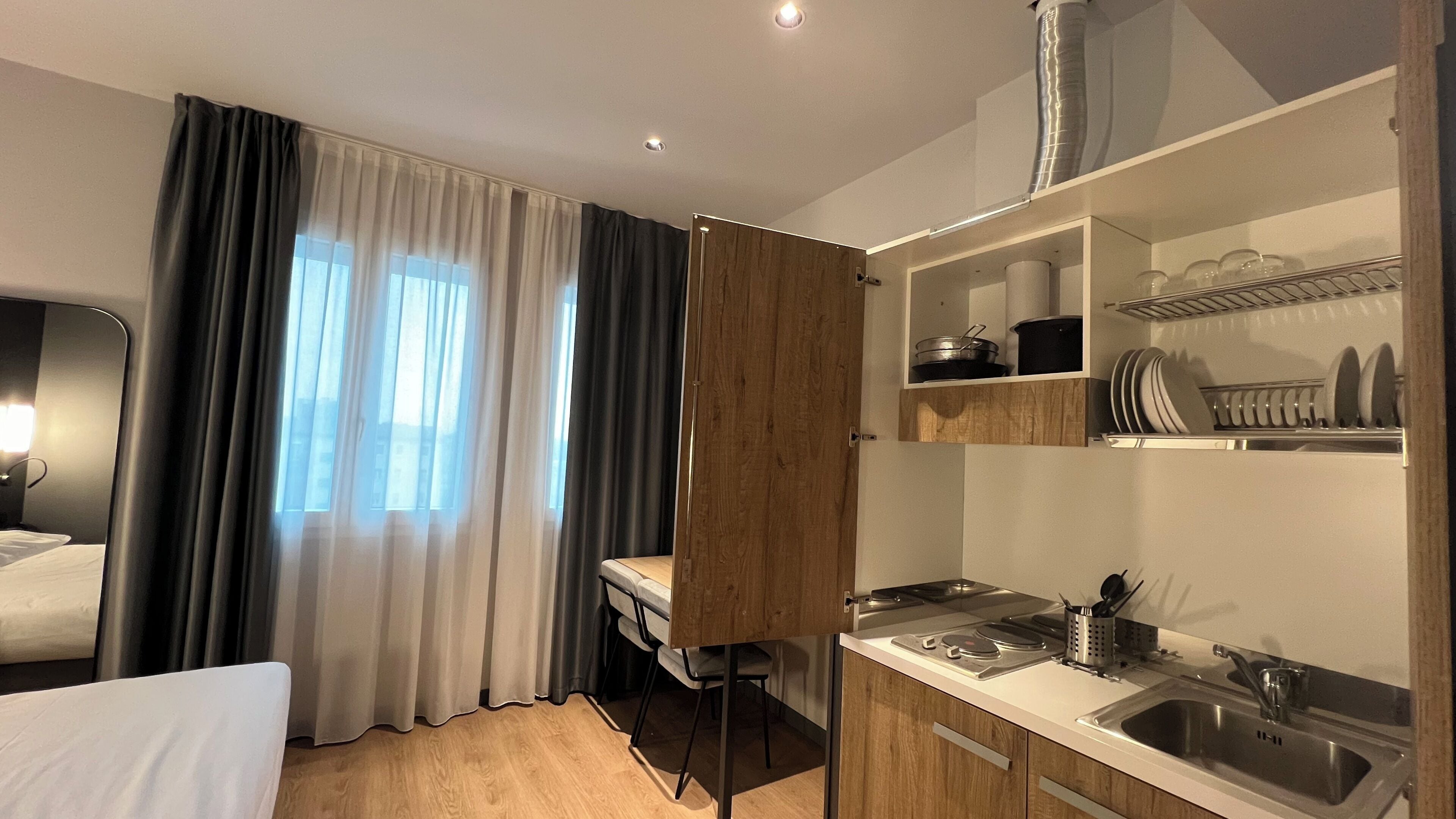 A compact hotel room with a bed, a small kitchenette, a wooden cabinet, and a window with curtains.