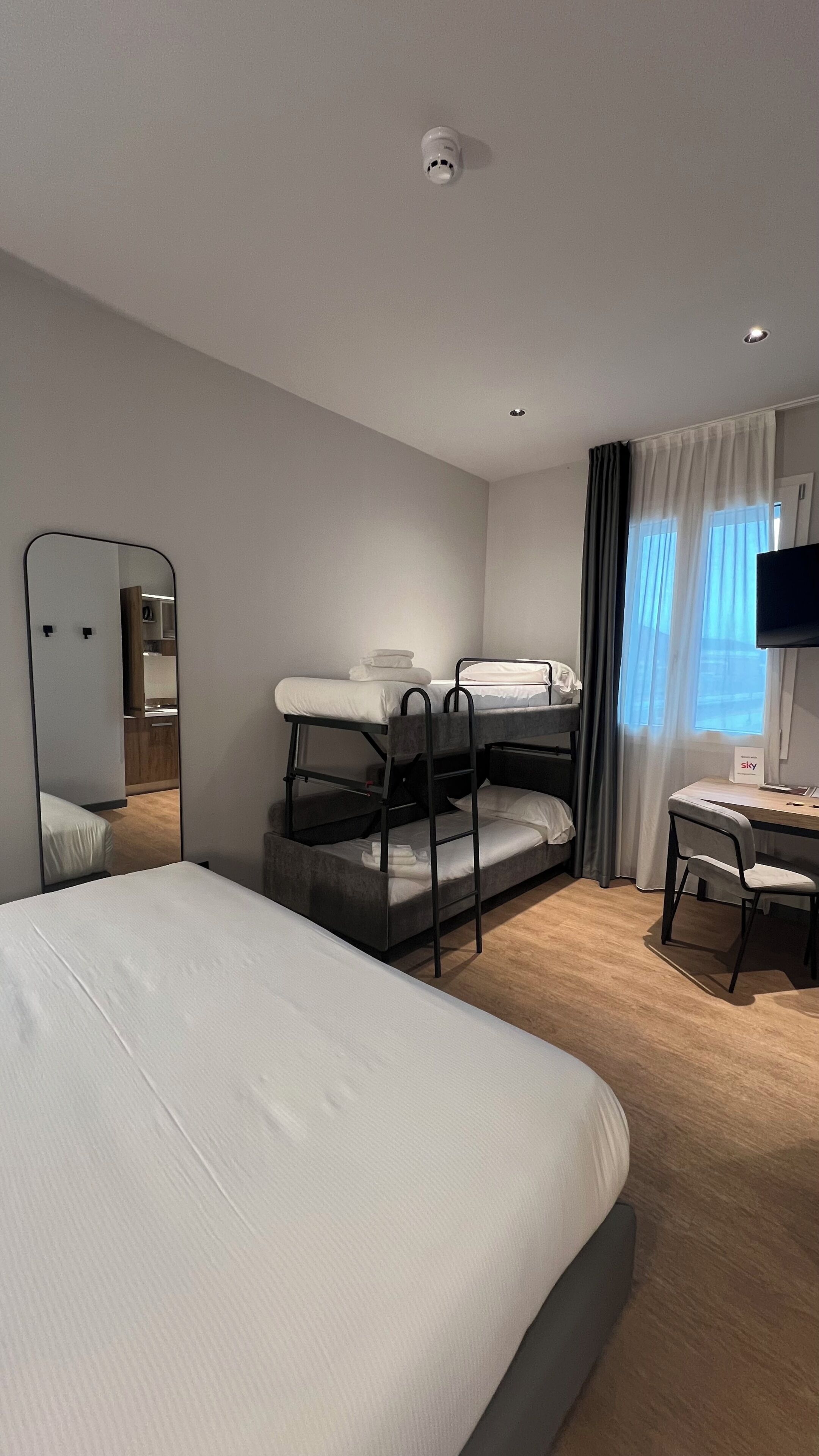 A hotel room with bunk beds, a desk, a chair, a flat-screen TV, and a mirror.