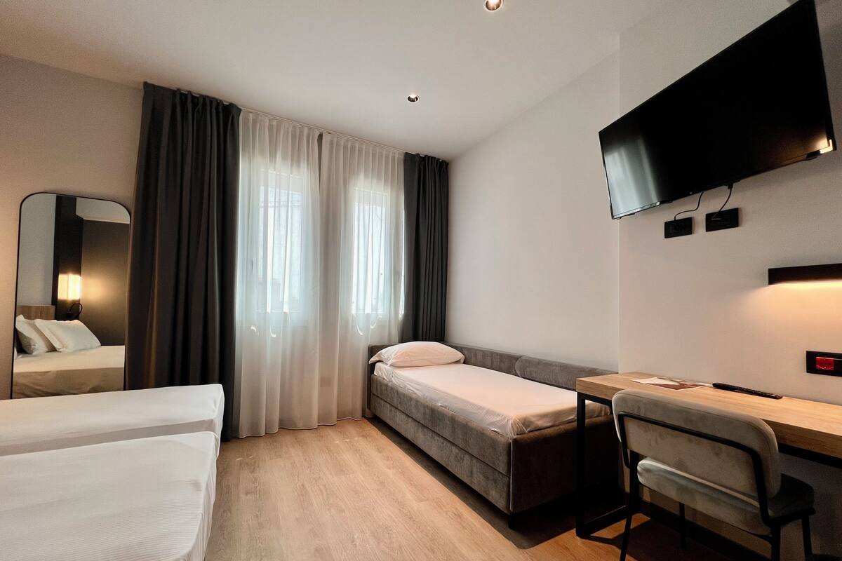 Una habitación de hotel con cama, escritorio, silla, televisor y ventana con cortinas.