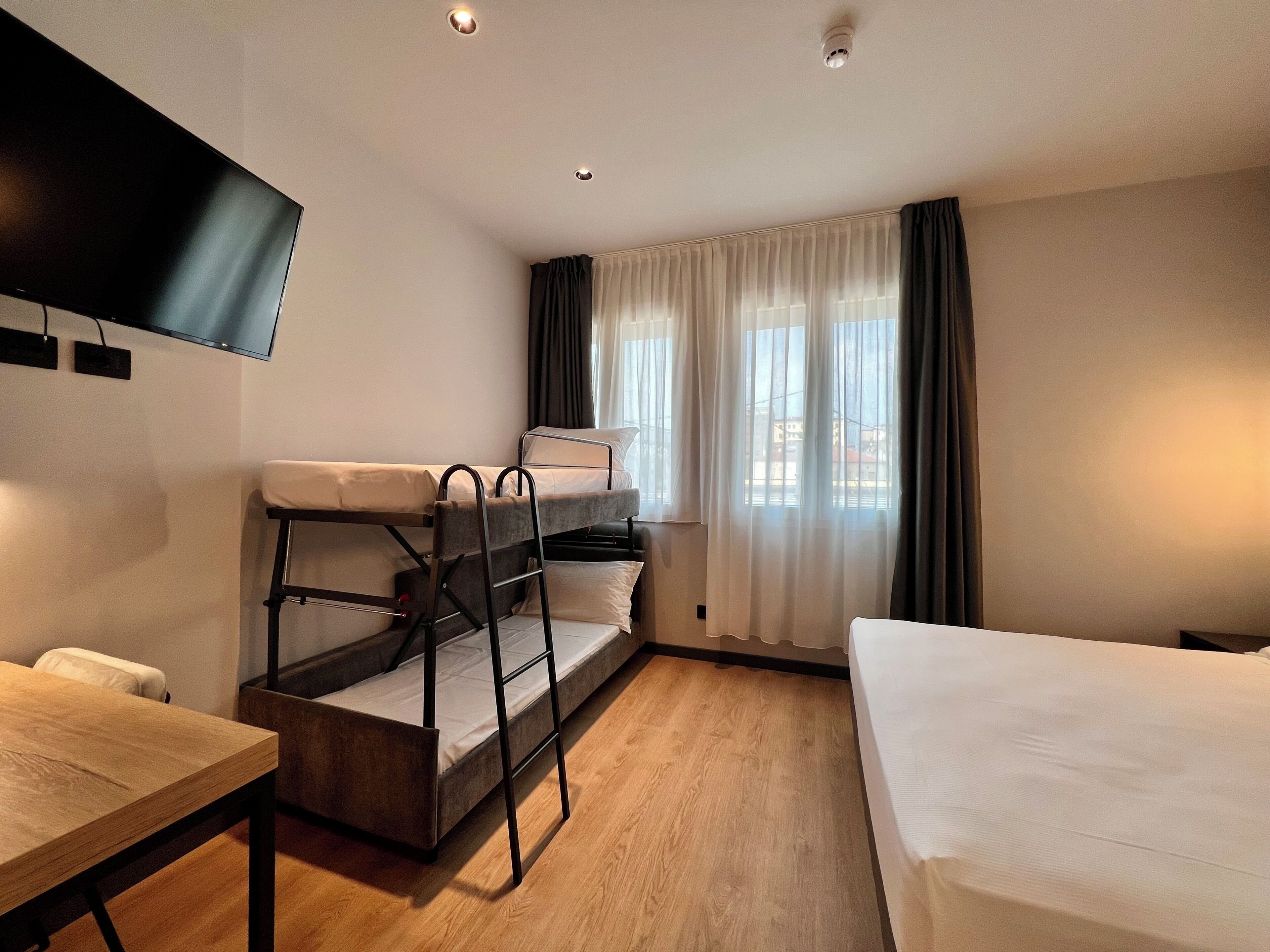 Habitación de hotel con literas, un televisor de pantalla plana, suelo de madera y una ventana con cortinas.