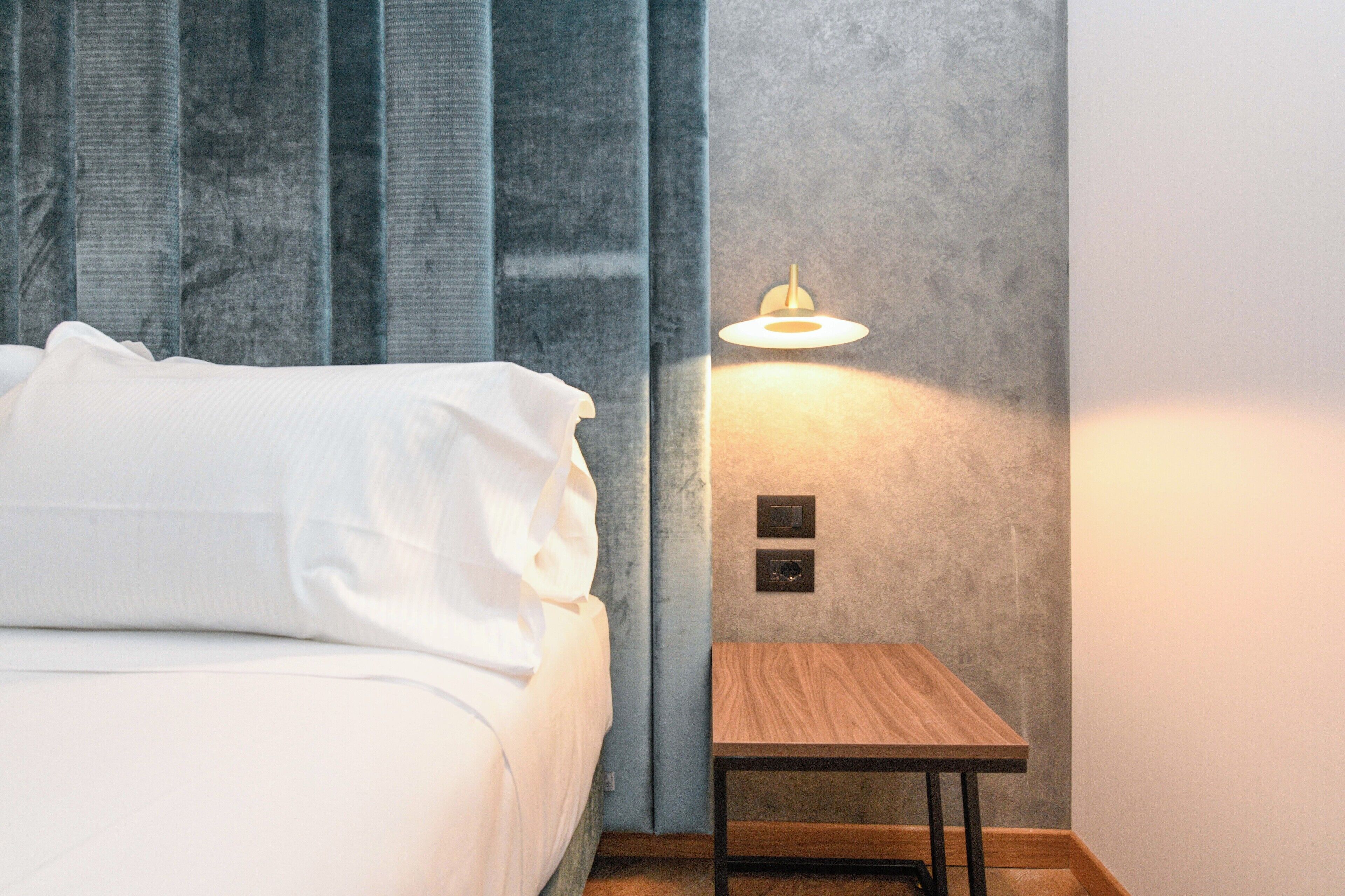 A neatly made bed with white linens, a small wooden nightstand with a lamp, and a textured grey wall.