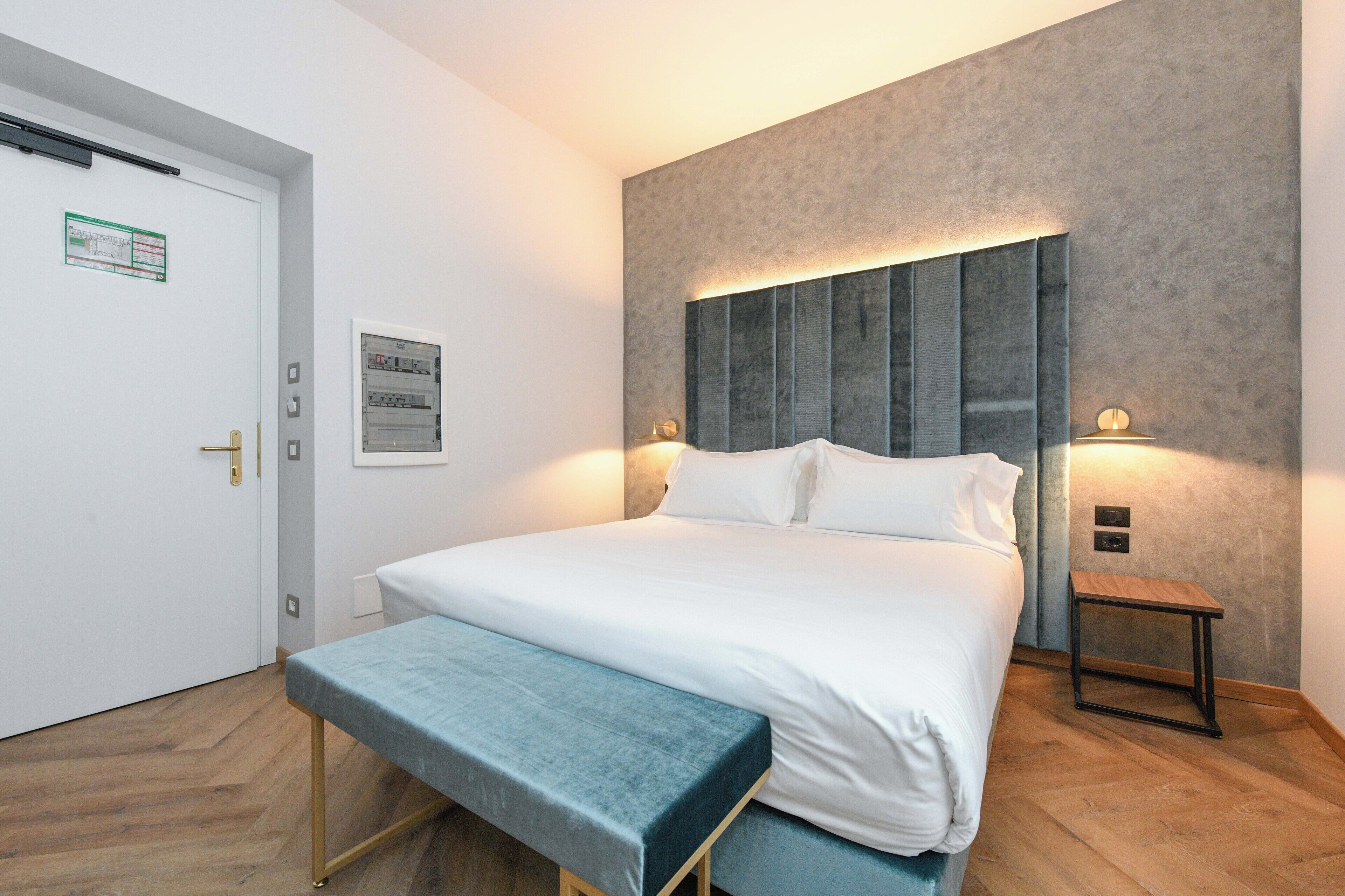 Una habitación de hotel moderna con una cama grande, un banco, una mesita pequeña y un aplique de pared.