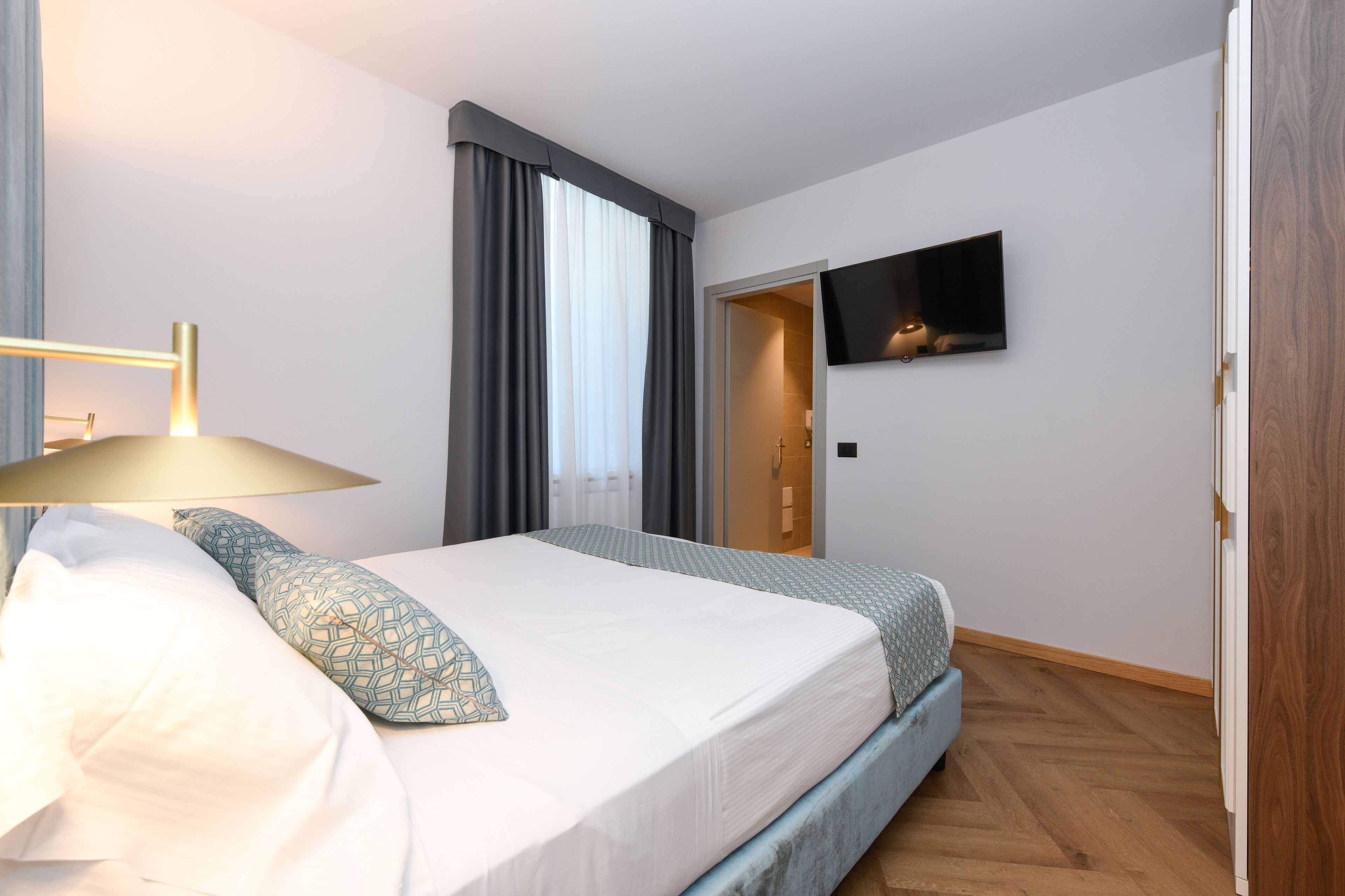 A neatly made bed with a patterned pillow, a bedside lamp, and a flat-screen TV mounted on the wall.