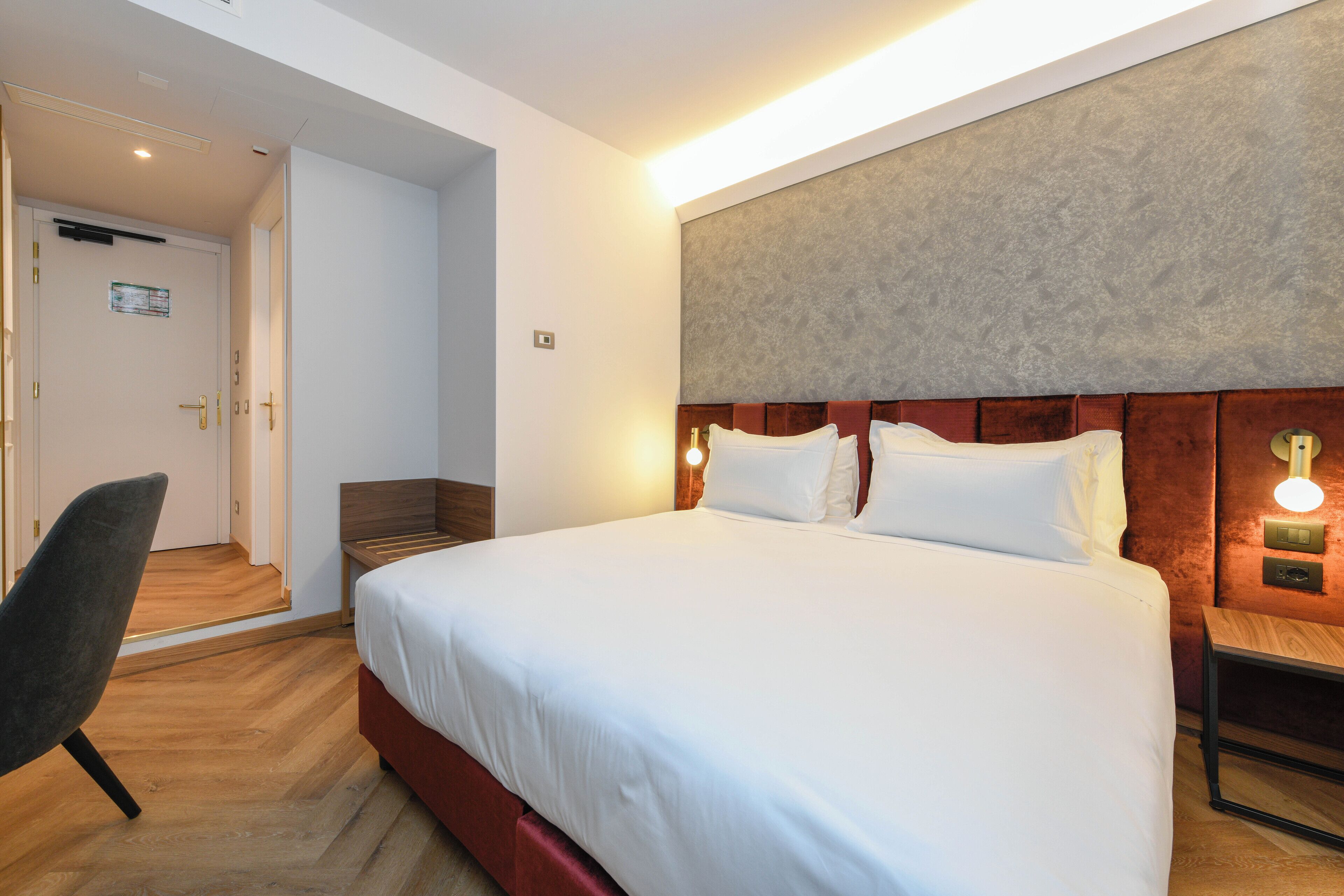 A hotel room with a large bed, a wooden nightstand, a chair, and a door leading to another room.