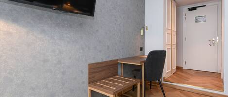 Une chambre moderne avec un bureau en bois, une chaise, un téléviseur fixé au mur et une porte donnant sur une autre pièce.