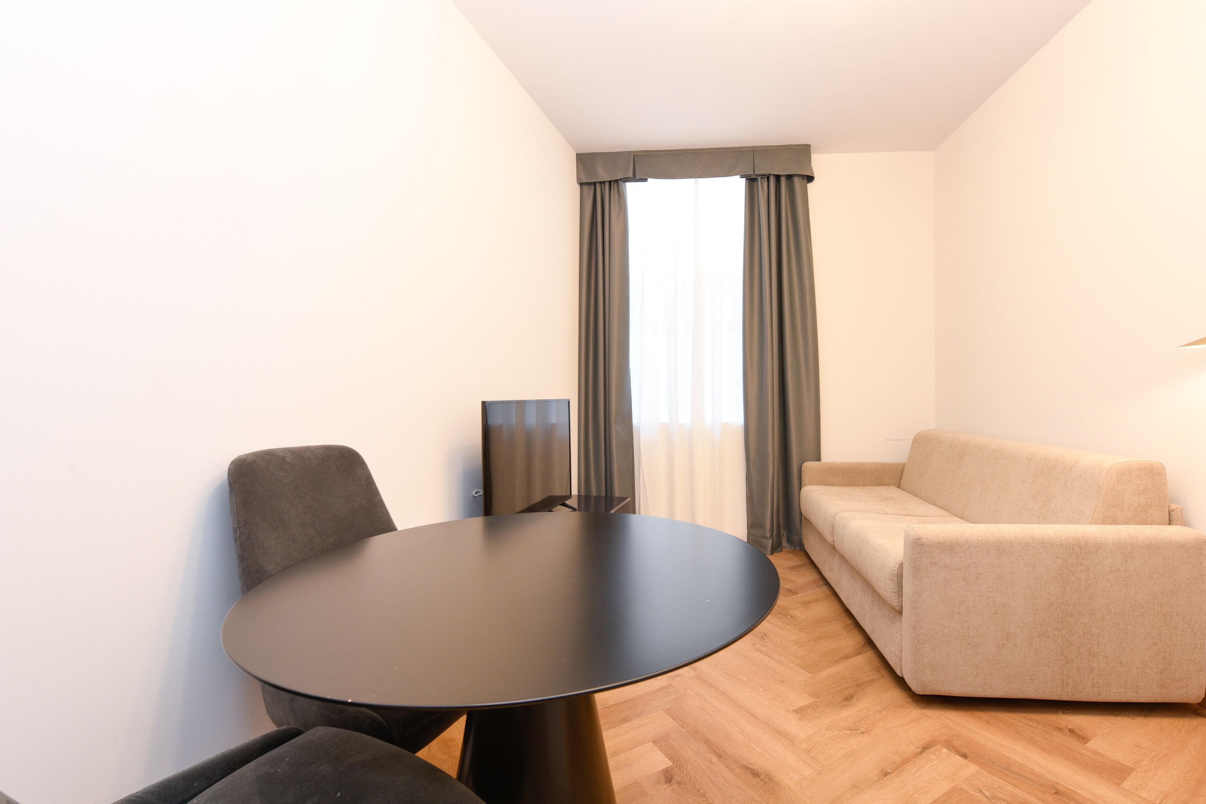 A modern room with a round black table, two grey chairs, a beige sofa, and a large window with grey curtains.