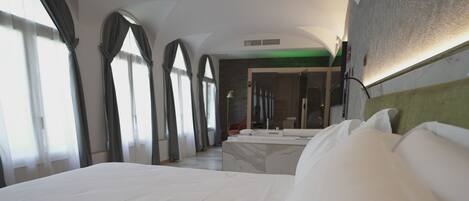 A modern hotel room with a large bed, a sitting area with a sofa and a small table, and a view of the hallway and another room.
