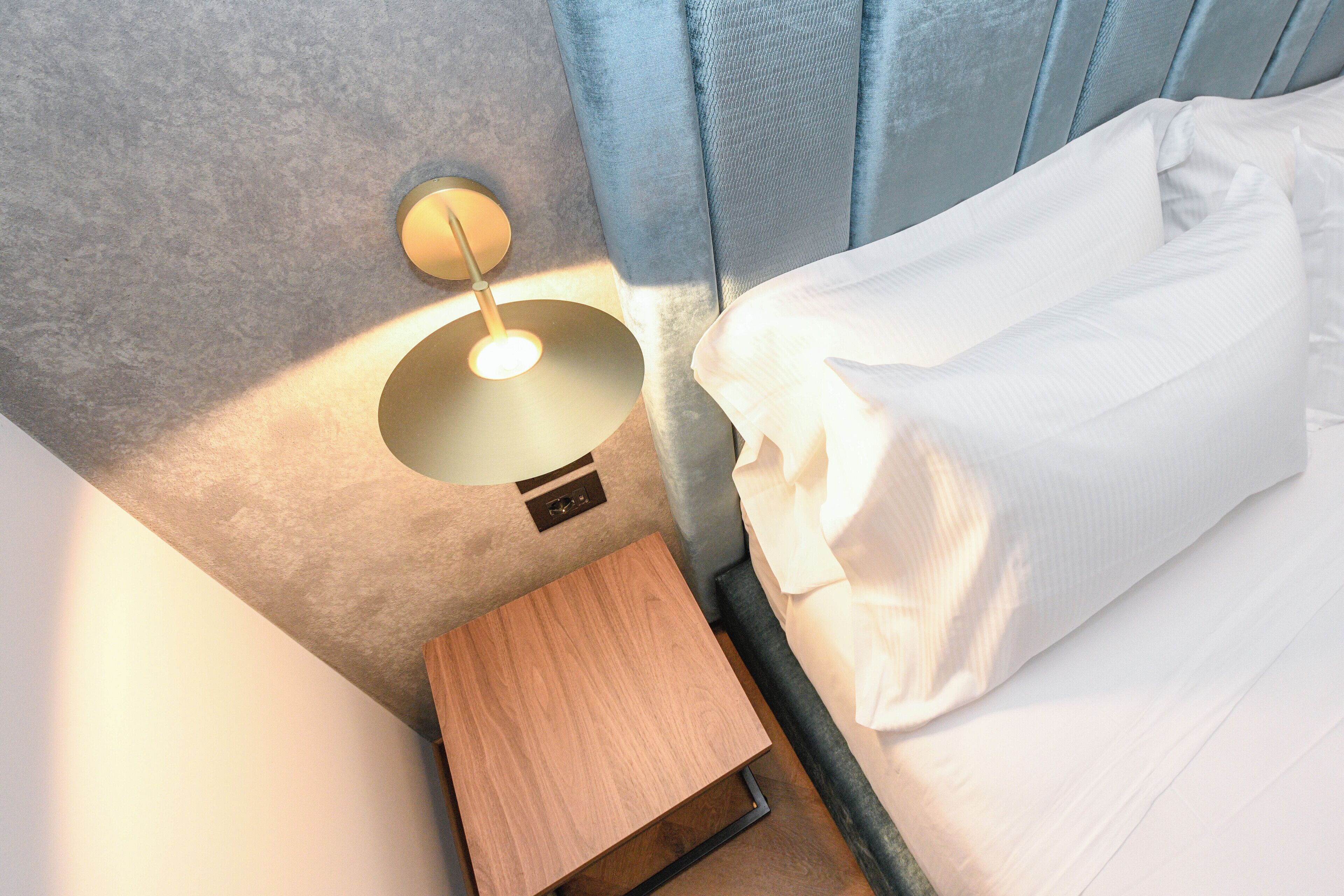 A neatly made bed with white linens, a bedside table with a lamp, and a blue curtain.