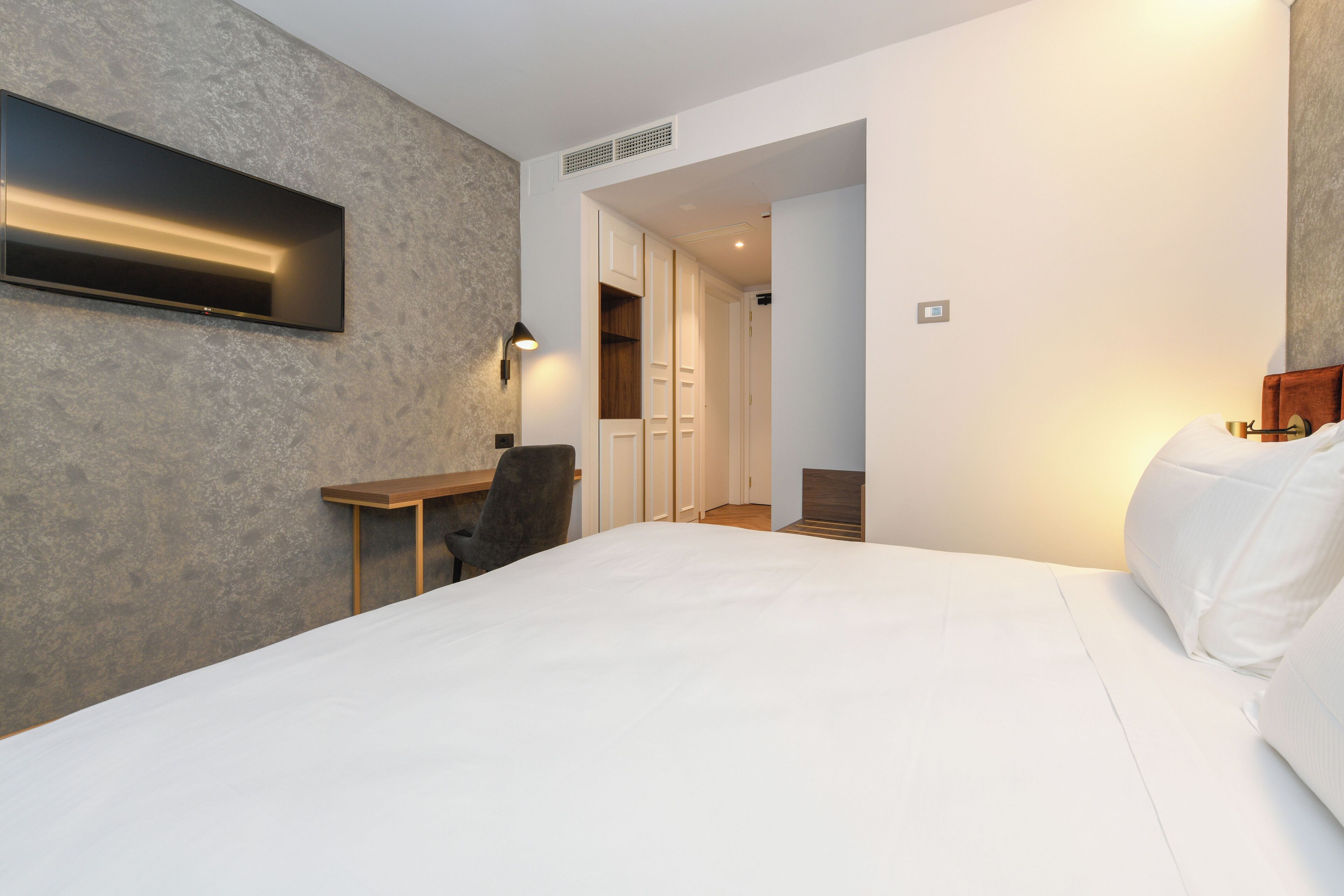 A hotel room with a large bed, a desk, and a chair. There is a wardrobe and a door leading to another room.