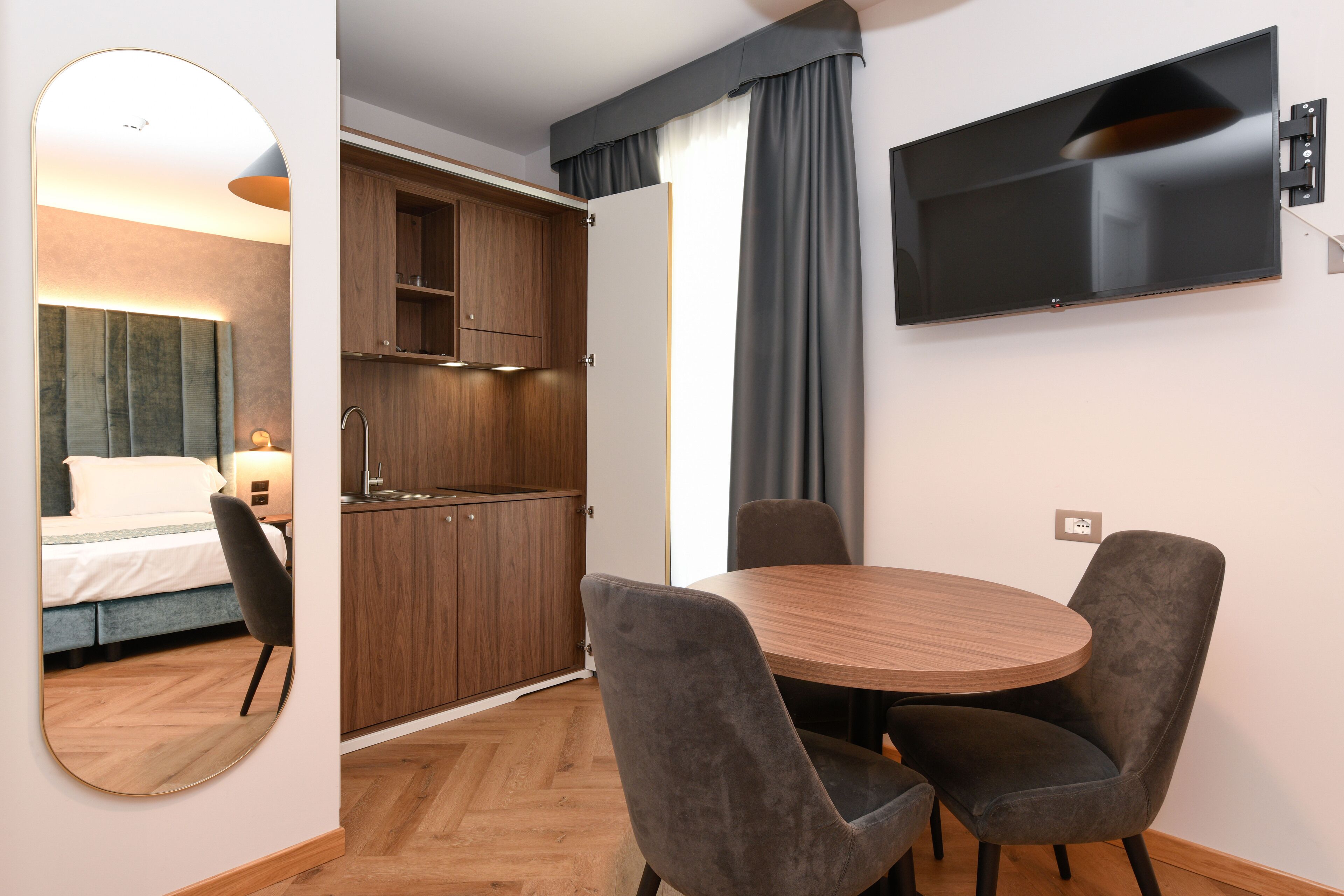 A hotel room with a bed, a round wooden table with four chairs, a TV mounted on the wall, and a wooden wardrobe.