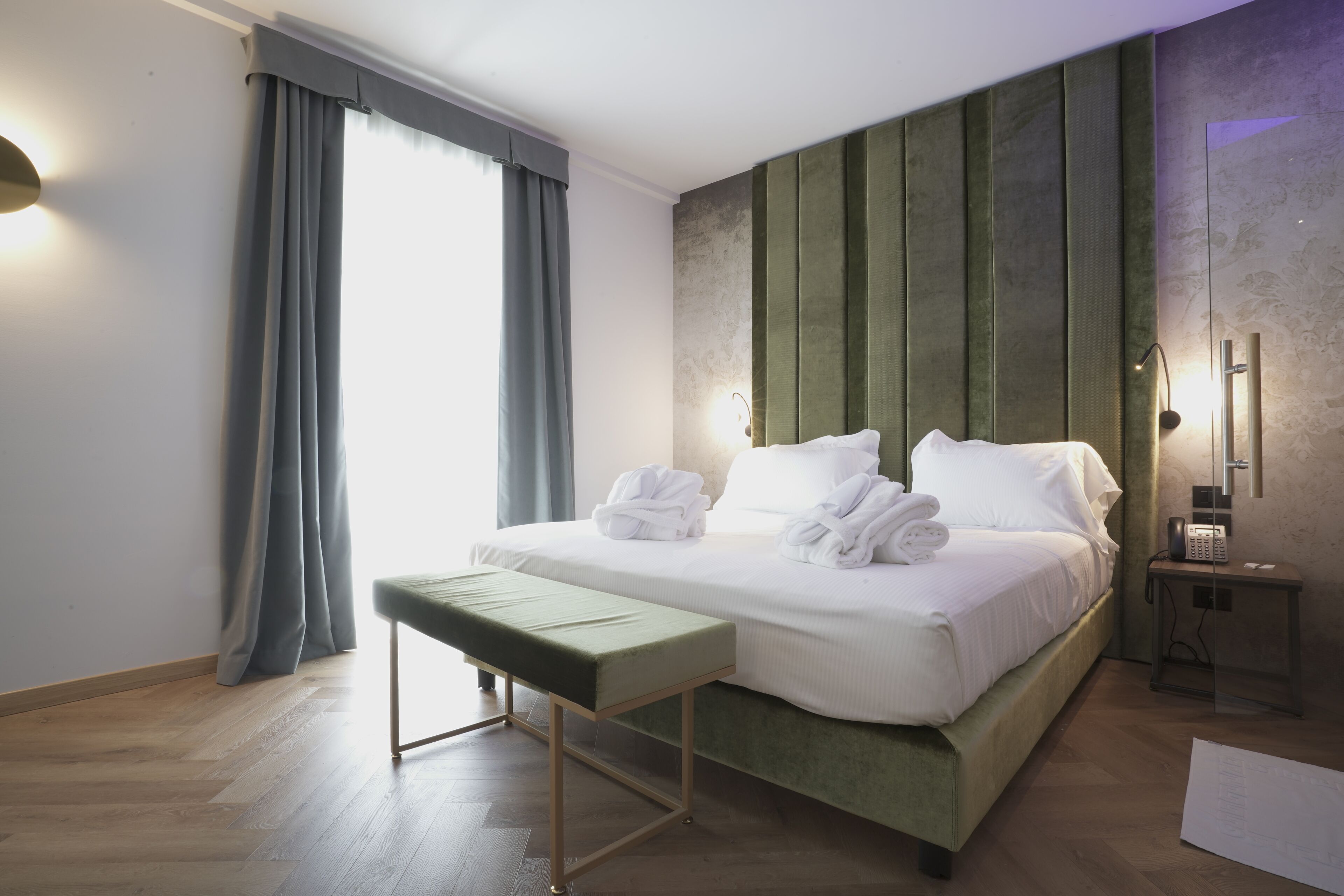A hotel room with a neatly made bed, a bench, a bedside table with a lamp, and a large window with curtains.