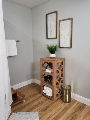 Rincón de una habitación con un estante de madera que sostiene toallas dobladas, una planta en maceta y un cuadro en la pared.