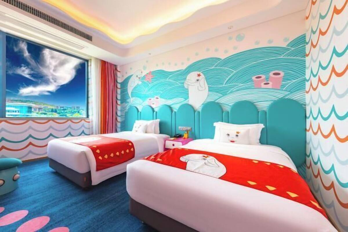 Habitación de hotel con dos camas, un ventanal amplio con vistas a la ciudad y un mural con criaturas marinas.