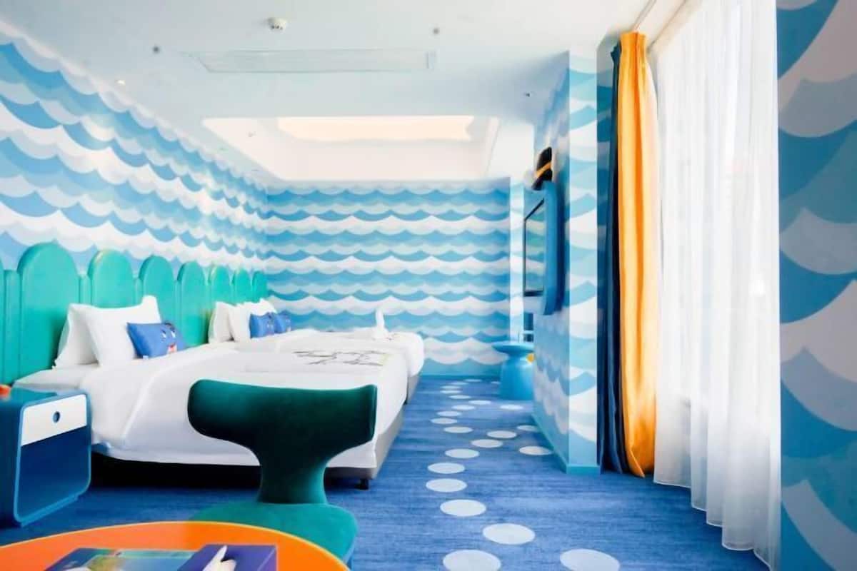 Una habitación de hotel con un papel pintado con diseño de olas, una cama con cabecera verde, una alfombra azul con lunares blancos y una ventana con cortinas de gasa.