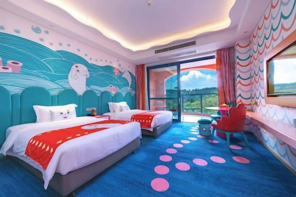 Una habitación de hotel con dos camas, un ventanal grande, un balcón y un mural colorido en la pared.