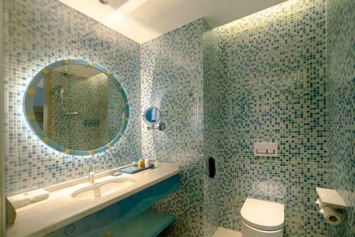 Un baño con paredes de mosaicos, un espejo redondo, un lavamanos y un inodoro.