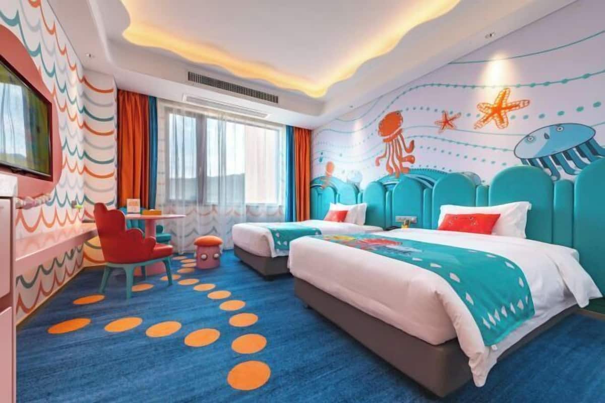 Habitación de hotel con dos camas, un televisor, un escritorio y un mural colorido en la pared.