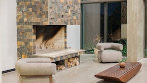 A modern living room with a stone fireplace, two cushioned chairs, a wooden coffee table, and a large window.