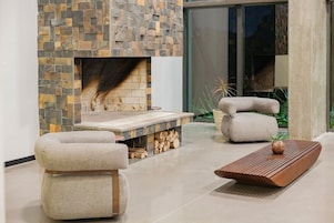 Uma sala de estar moderna com uma lareira de pedra, duas poltronas acolchoadas, uma mesa de centro de madeira e uma janela ampla.