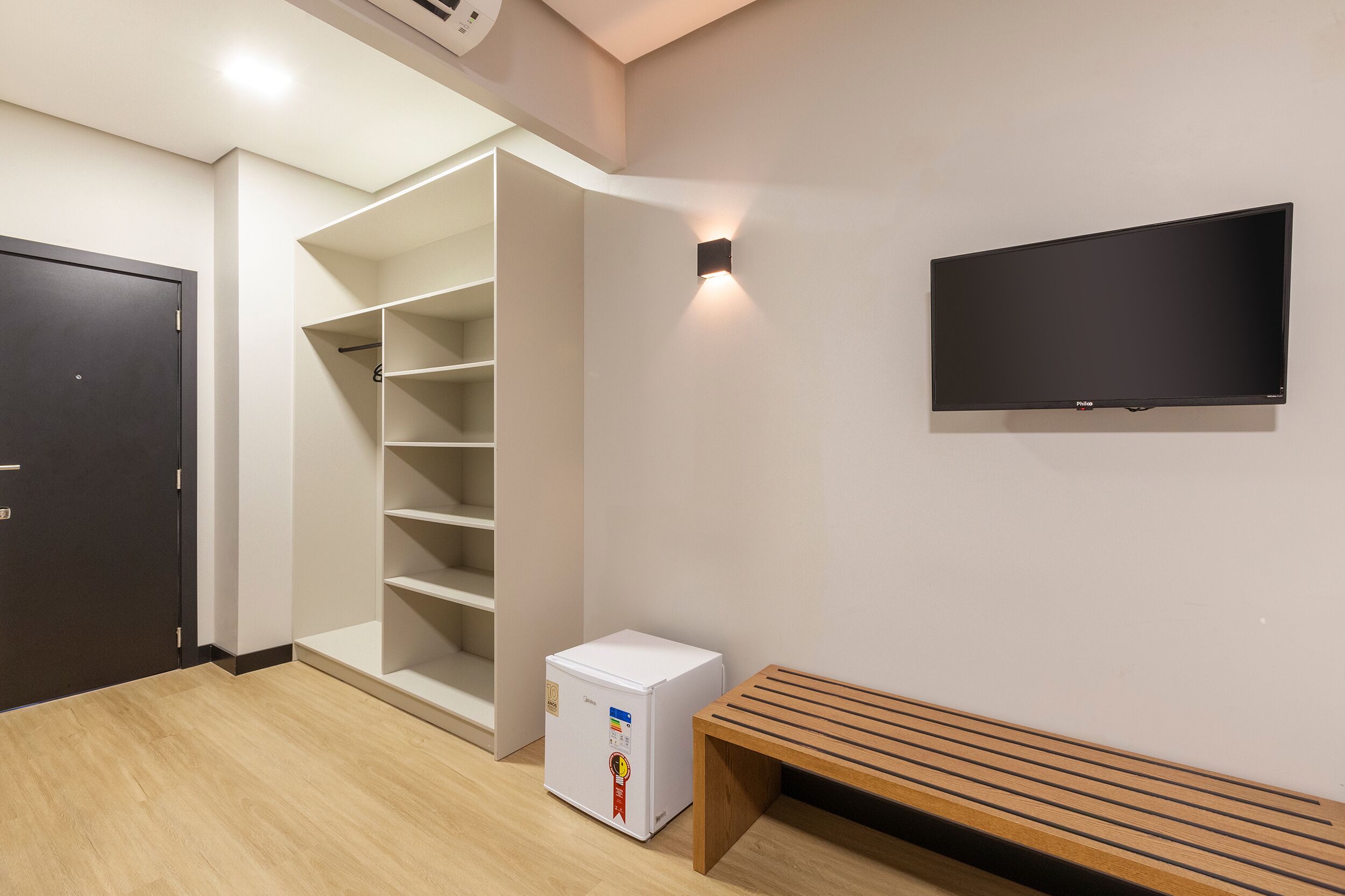 A room with a white wardrobe, a white mini-fridge, a wooden bench, and a mounted TV.