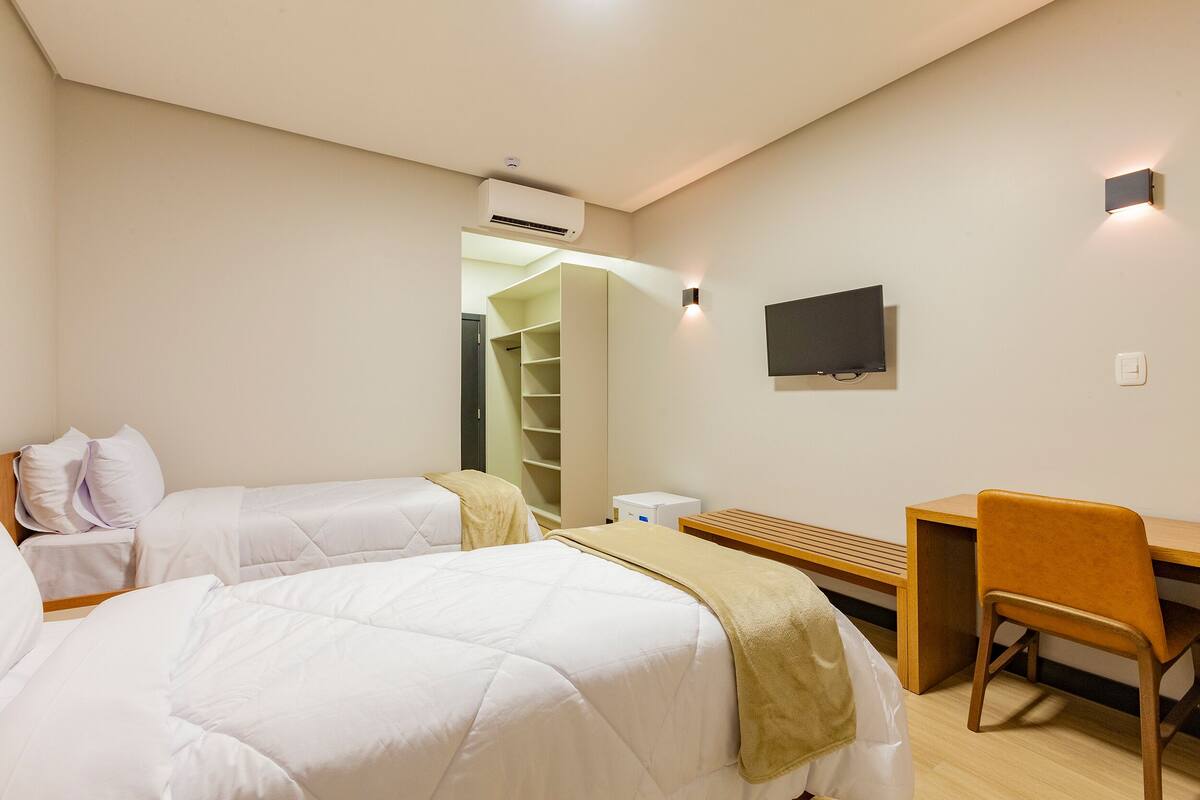 Habitación de hotel con dos camas individuales, un escritorio, una silla, un televisor y un armario.