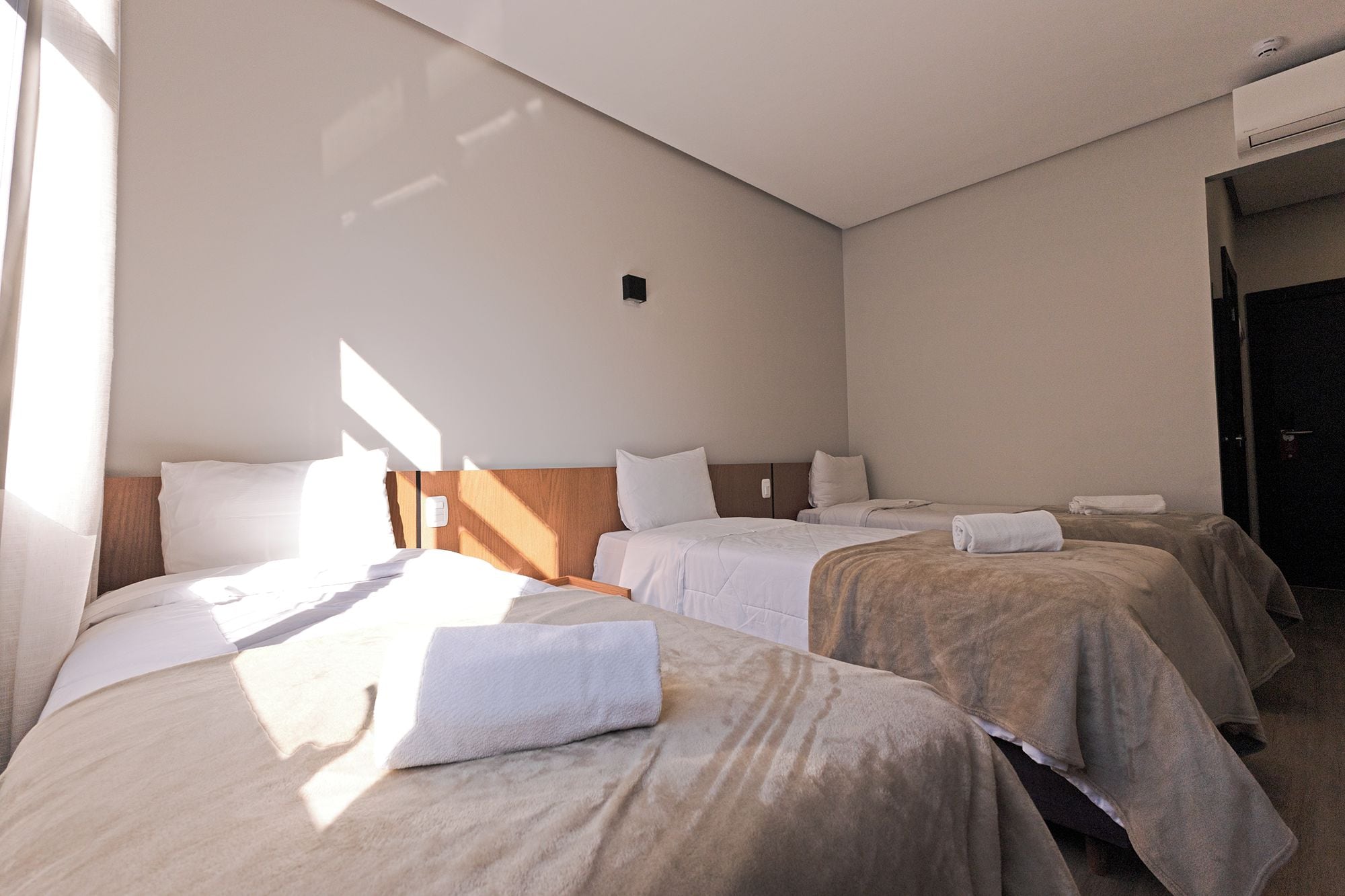 Ett hotellrum med två sängar, var och en med en vit kudde och ett brunt täcke. Det finns ett fönster som släpper in naturligt ljus, och en luftkonditioneringsanläggning som är monterad på väggen.