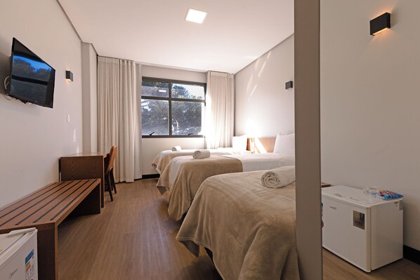 A hotel room with a bed, bedside table, a desk, a chair, a TV mounted on the wall, and a window with curtains.
