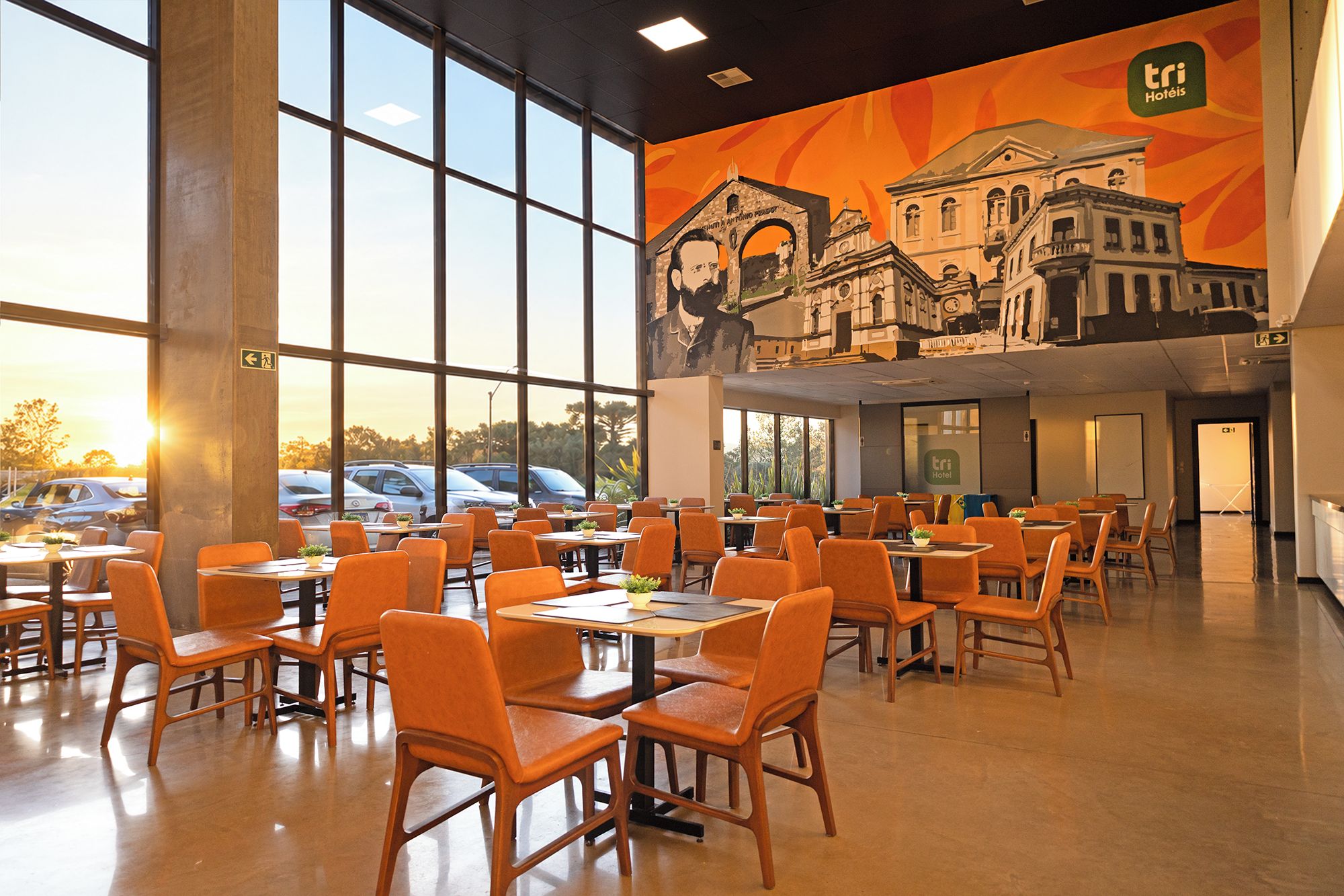 Un comedor amplio e interior con varias mesas y sillas, ventanas grandes y un mural de un edificio en la pared.