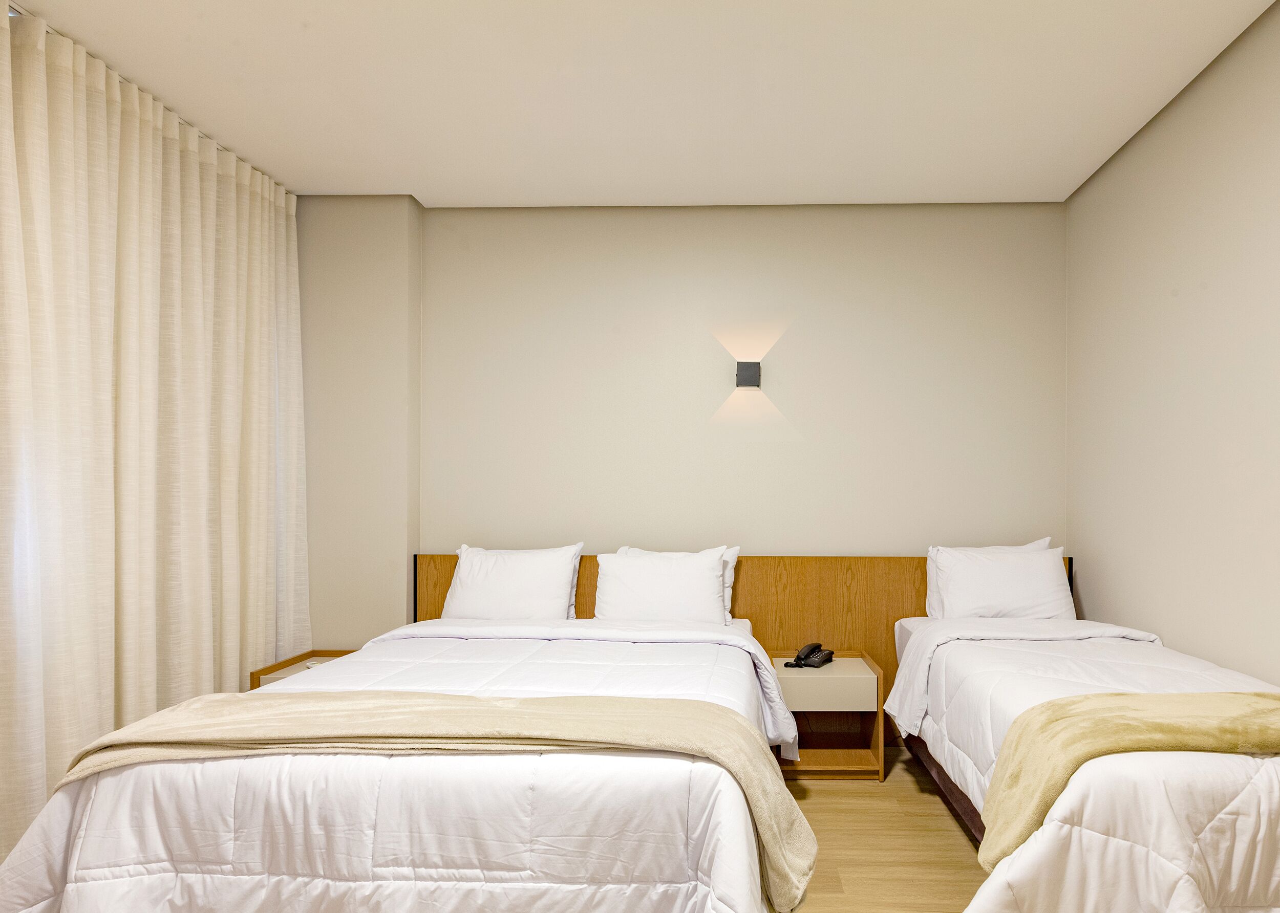 A hotel room with two beds, a wooden headboard, a bedside table with a phone, and a wall-mounted light fixture.