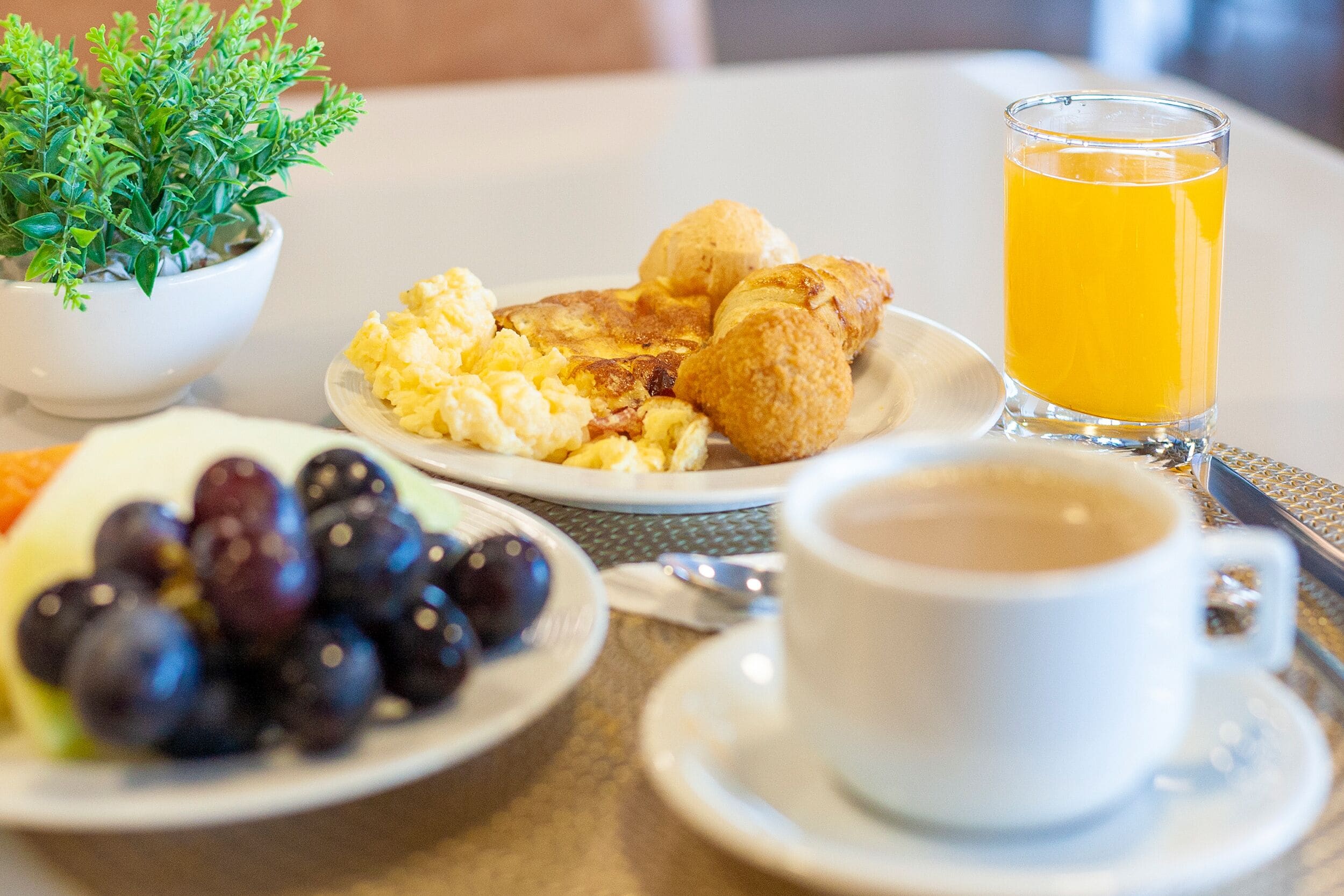 A breakfast setting with scrambled eggs, croissants, a glass of orange juice, a cup of coffee, and a bowl of grapes on a table.