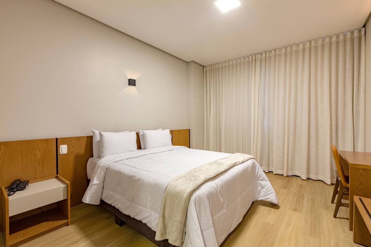Habitación de hotel con cama, mesita de noche con teléfono, escritorio y una silla, y una cortina.