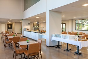 Uma sala de jantar ampla com várias mesas e cadeiras, um balcão de buffet com diversos alimentos e vista para o exterior através de grandes janelas.