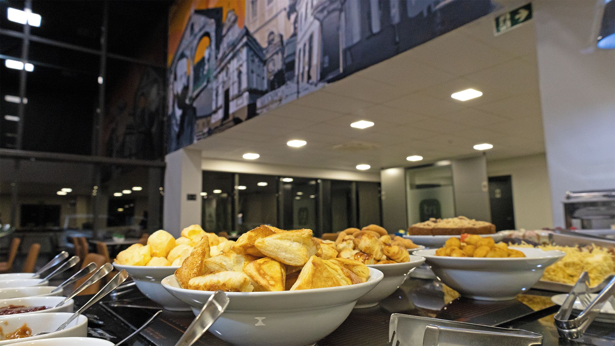 A buffet with various dishes, including bread and pastries, in a modern dining area with a mural on the wall.