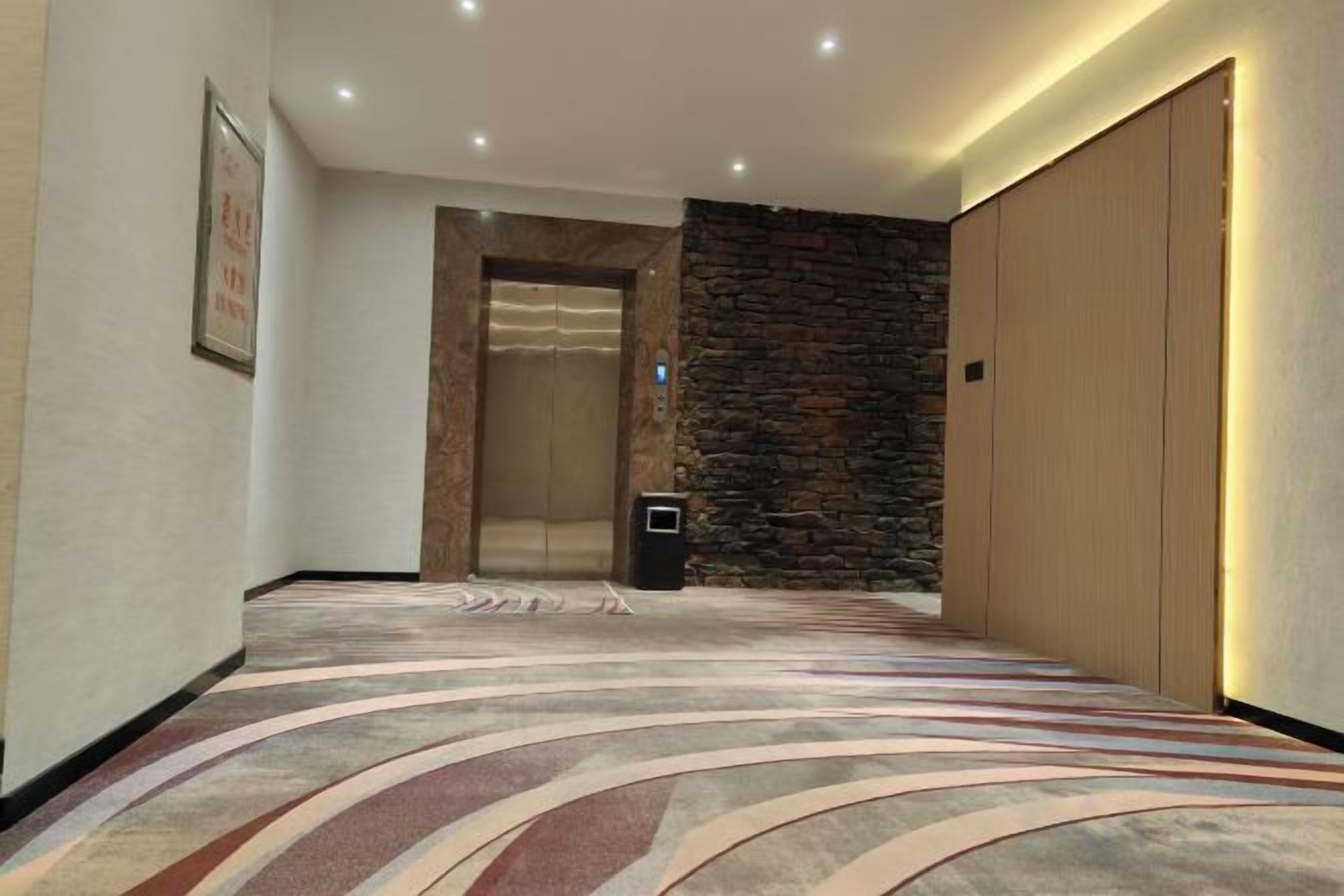 A hotel corridor with a patterned carpet, an elevator, a stone wall, and a closed door.