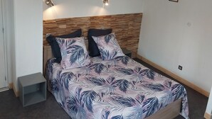 A bedroom with a wooden headboard, a bed with a leaf-patterned bedspread, two wall-mounted lights, and a small bedside table.