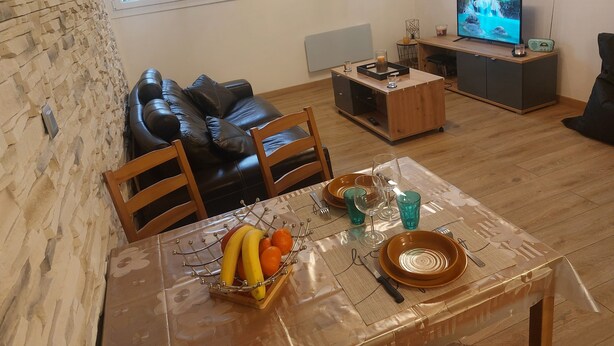 A living room with a stone wall, a black leather sofa, a wooden dining table set with a fruit bowl, and a television on a wooden cabinet.