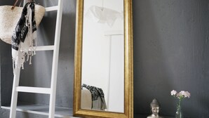 Un espejo con marco dorado, una escalera blanca, un bolso tejido, una estatua de Buda y un jarrón con flores sobre una repisa frente a una pared gris.