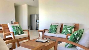 A modern living room with wooden furniture and white cushions, a small table, and a visible kitchen area in the background. - Playa Caracol Residences 1 and Beach Club Chame Banks (La Boca de Chame)