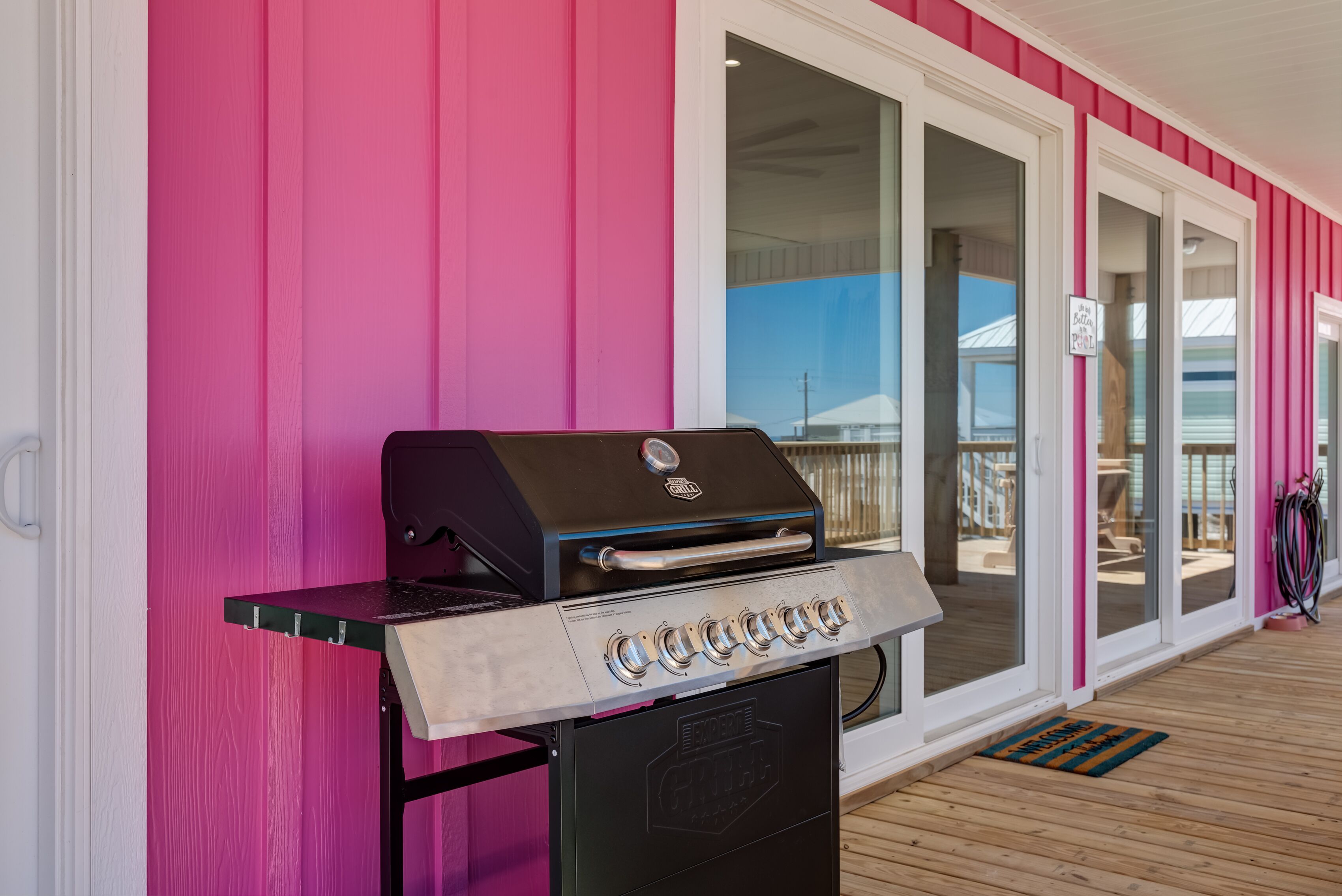 Ein Grill mit mehreren Brennern auf einer Holzterrasse neben einem Gebäude mit rosa Fassade und großen Glasfenstern.