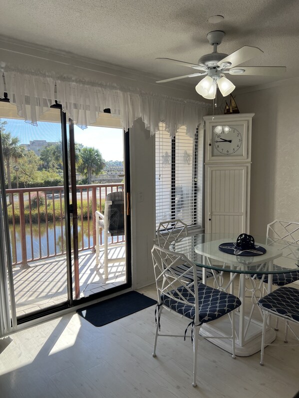 A room with a glass table and chairs, a ceiling fan, a clock, and a view of a body of water through a sliding glass door.