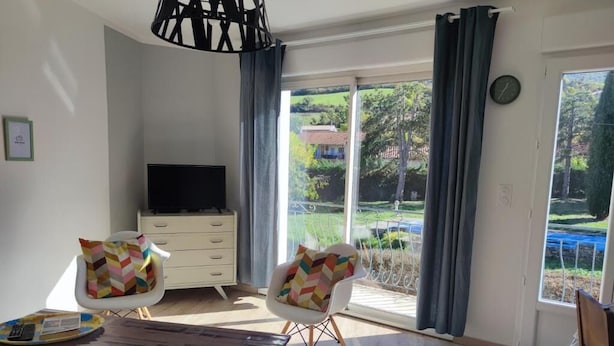 A modern living room with a TV, two chairs with colorful cushions, a wooden coffee table, and a large sliding glass door leading to a garden. - 2 Bedroom Apartment in Saint-Georges-de-LuzenÃ§on (Saint-Georges-de-Luzençon)
