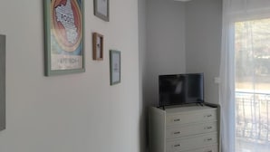 A room with a TV on a white dresser, a balcony with a white curtain, and several framed pictures on the wall. - 2 Bedroom Apartment in Saint-Georges-de-LuzenÃ§on (Saint-Georges-de-Luzençon)