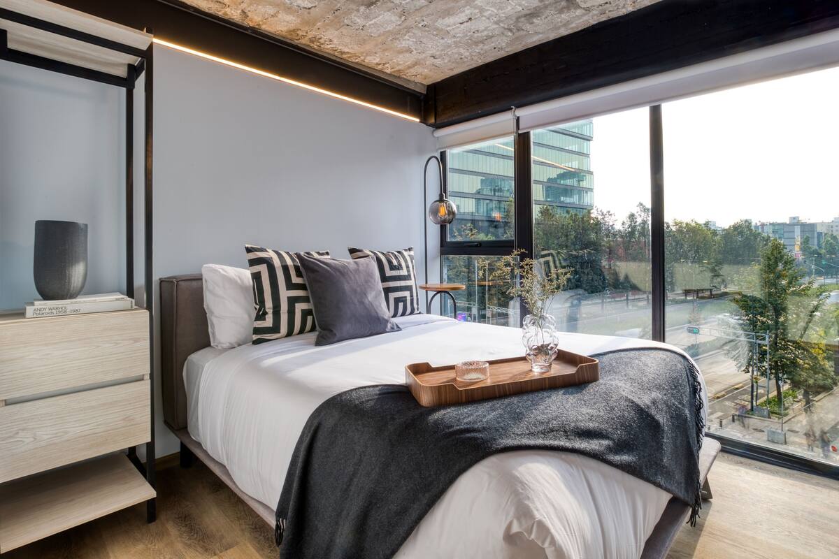 A modern bedroom with a large bed, a bedside table, a window with a city view, and a wooden tray on the bed.