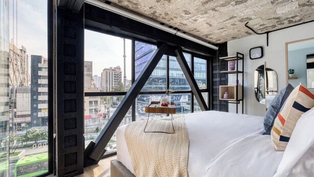 A modern hotel room with a large bed, a view of the cityscape, and a concrete ceiling. - ULIV El Ángel (Mexico City)
