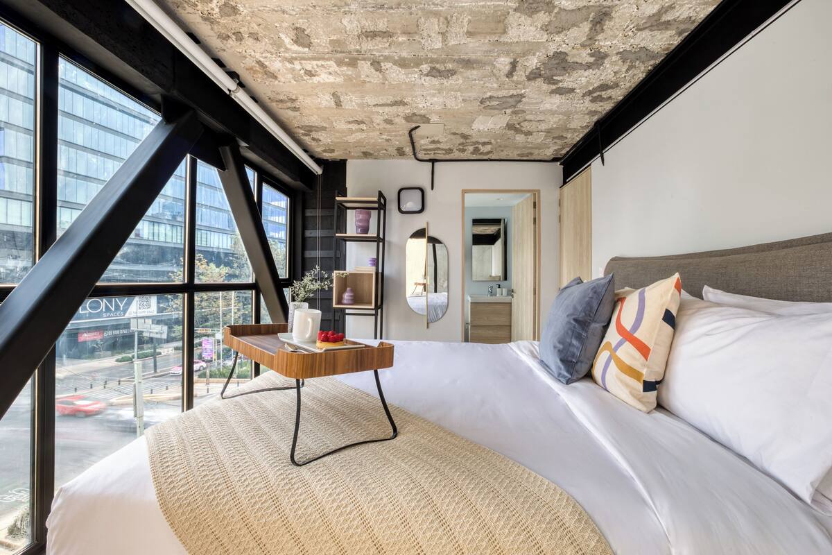 A modern hotel room with a large bed, a small wooden side table, a view of a cityscape through large windows, and a concrete ceiling.