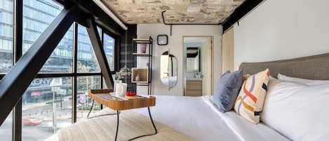 A modern hotel room with a large bed, a small wooden side table, a view of a cityscape through large windows, and a concrete ceiling.