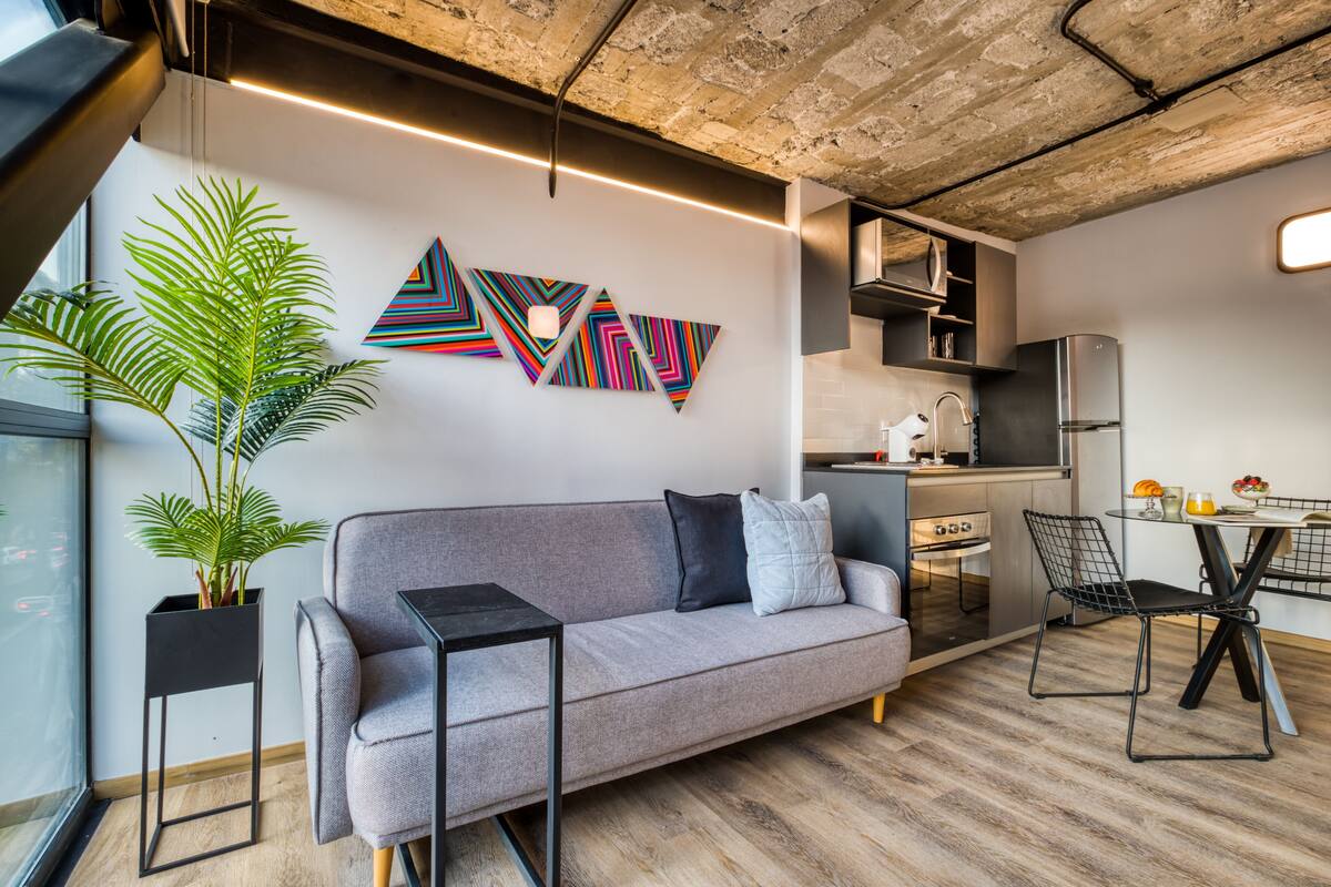 A modern living space with a grey sofa, a black side table, a potted plant, and a kitchen area with a refrigerator and a dining table with chairs.