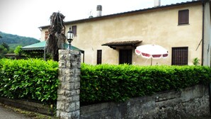 A building with a stone pillar, a green umbrella, and a hedge in front. - La Cheta - House in the hills with garden, between Lucca and the beaches of Viareggio (Orbicciano, Camaiore)