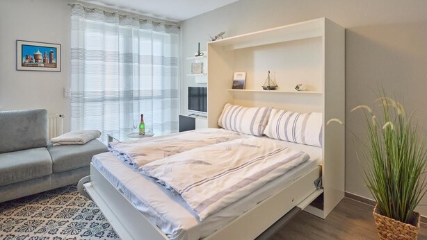 Een slaapkamer met een bed, een bank, een nachtkastje, een lamp, een raam met gordijnen, een wandplank met decoratieve objecten, en een potplant.